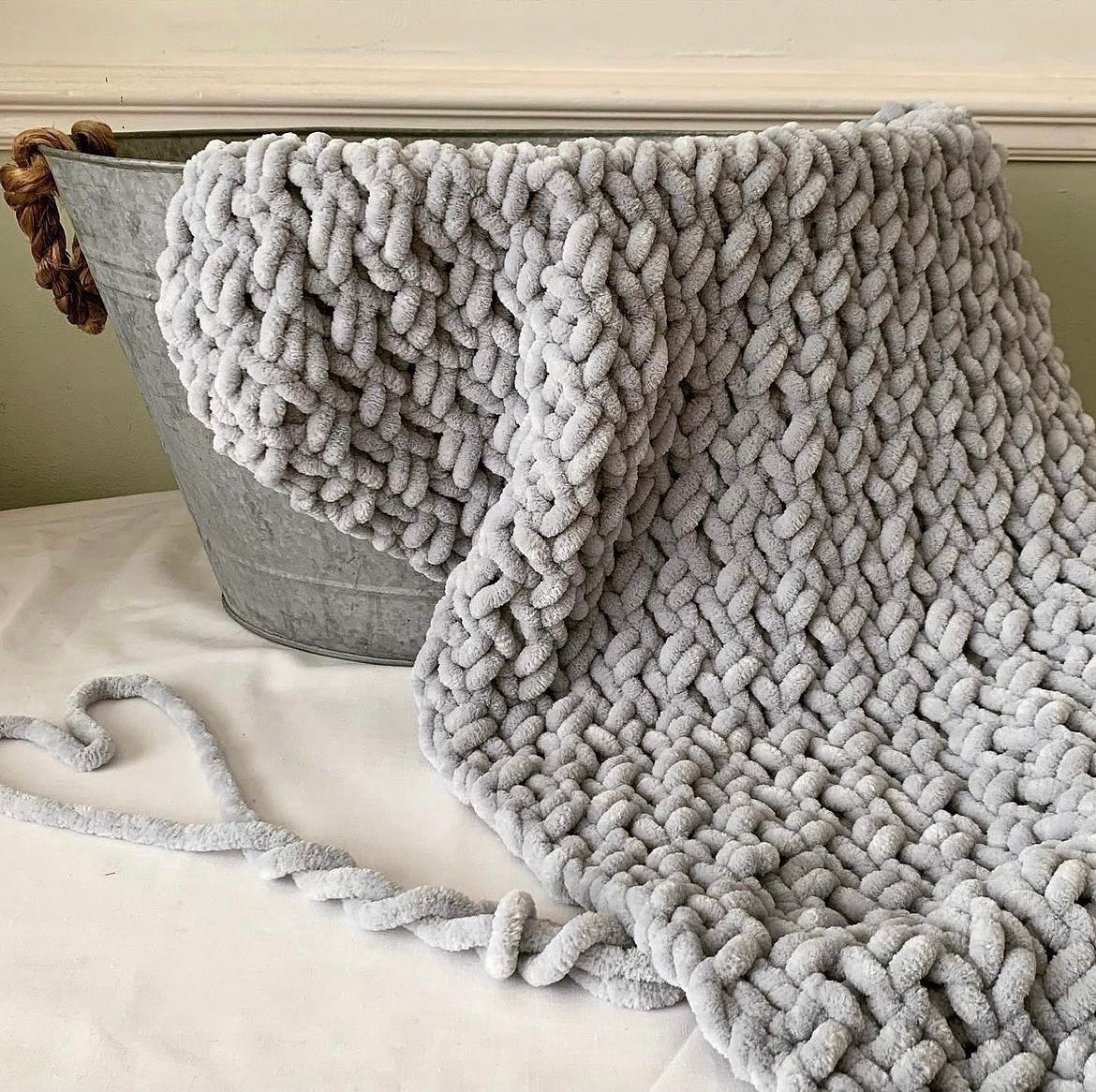 Hand-knitted silver grey chenille baby blanket draped over a metal rustic basket, featuring a thick, soft, chunky knit pattern—perfect for cosy nursery decor or newborn gifts.