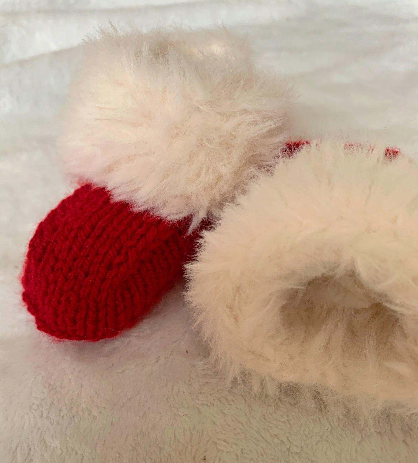 Close-up of SnugaBug’s Christmas SnUGG Boots—red knitted baby booties with fluffy white cuffs—resting on a soft textured surface. Perfect for gifting and featured in our Fiver Friday Deal.