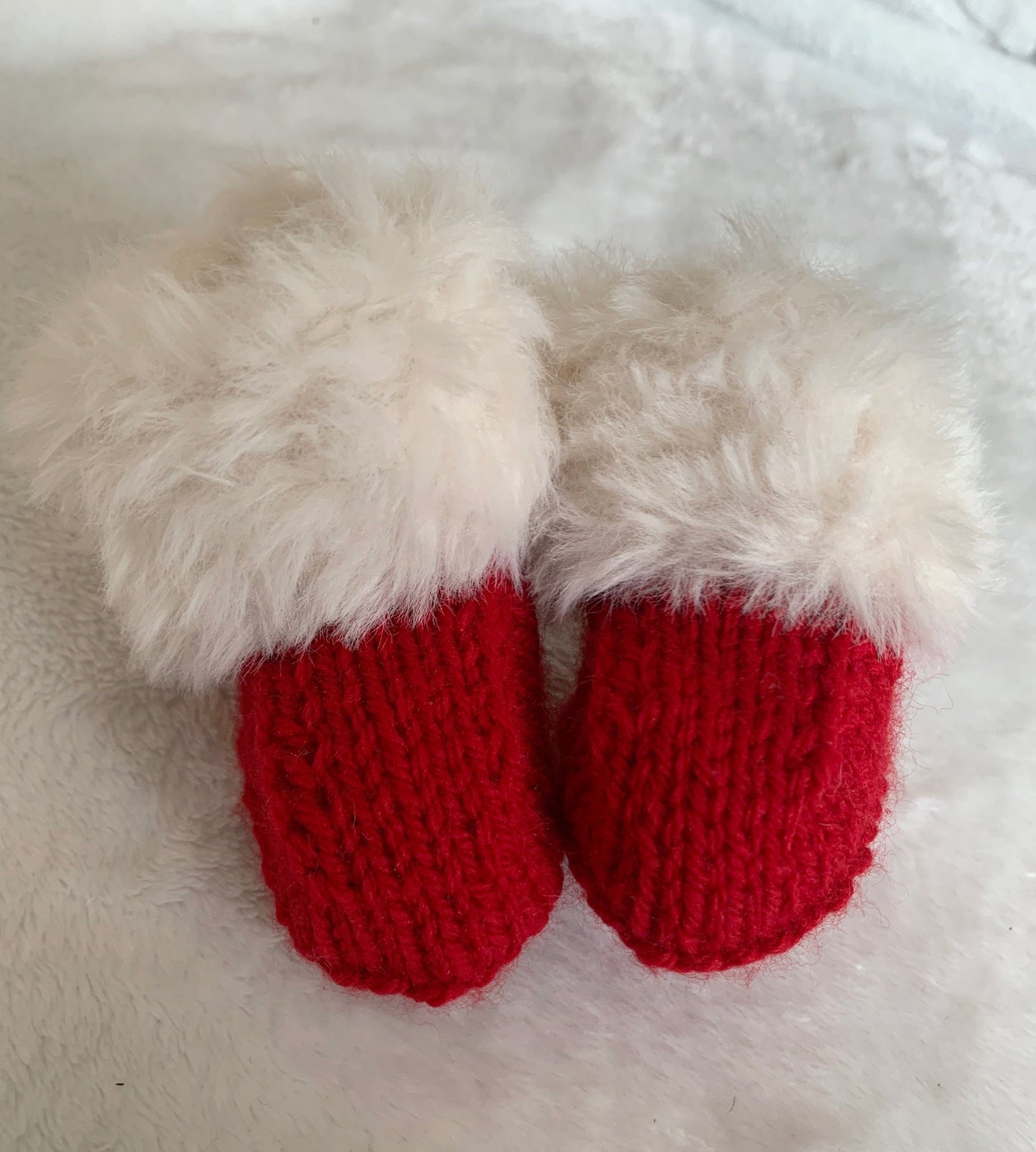 SnugaBug Christmas SnUGG Boots: small red knitted baby booties with fluffy white cuffs, Santa style, on soft white fabric—a perfect gift and part of our Fiver Friday Deal.