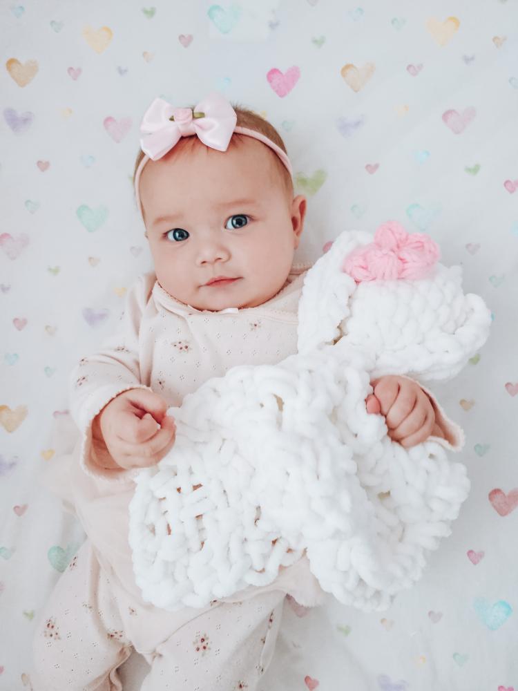 Baby girl in pink outfit and bow headband holding a white hand-knitted chunky chenille bunny lovey comforter with pink puff flower by SnugaBug