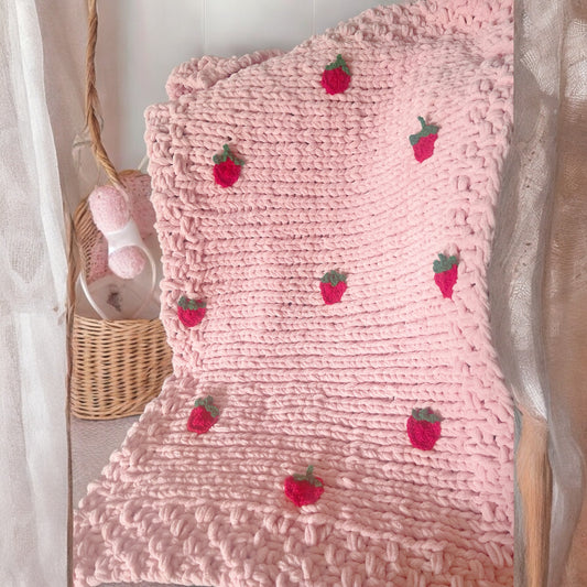 A dusky pink baby blanket with red crochet strawberries on it, displayed on a bed with a basket and a teddy bear in the background.