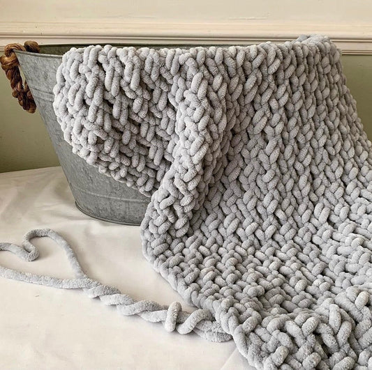 Hand-knitted gray chenille baby blanket draped over a metal basket, featuring a thick, soft, chunky knit pattern—perfect for cozy nursery decor or newborn gifts.