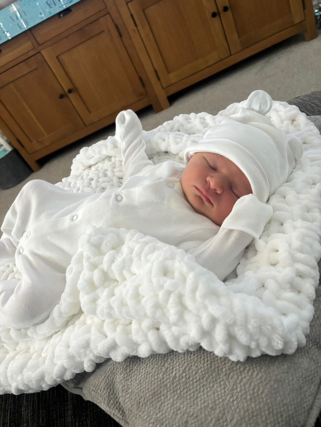 Newborn baby wrapped in a white chenille blanket with a matching hat, lying on a textured surface.
